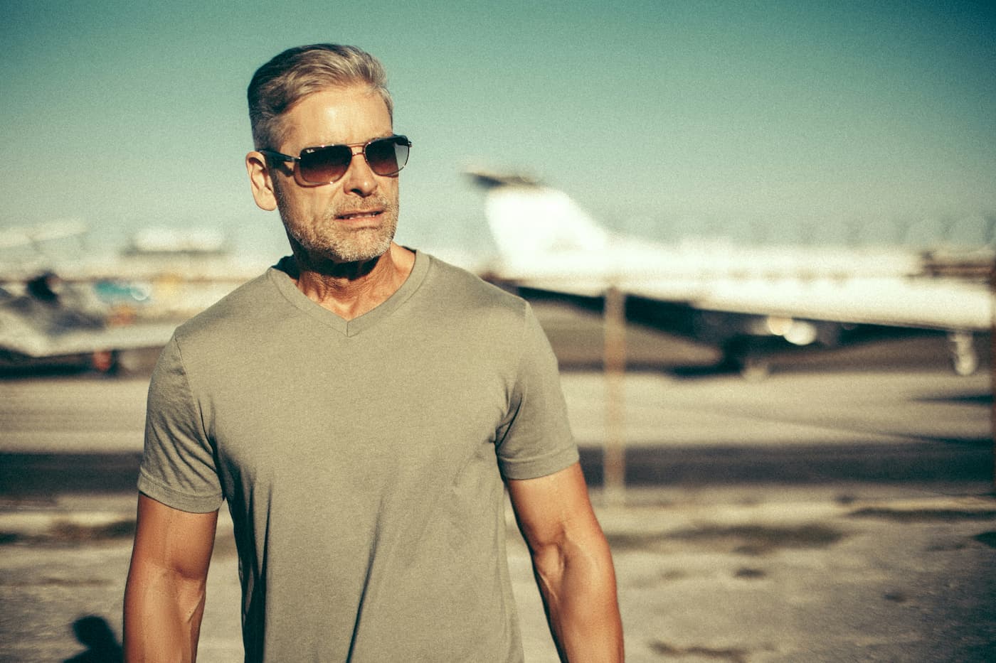 Confident middle-aged man with gray hair wearing sunglasses and a fitted gray T-shirt outdoors, representing men's health, vitality, and healthy aging.