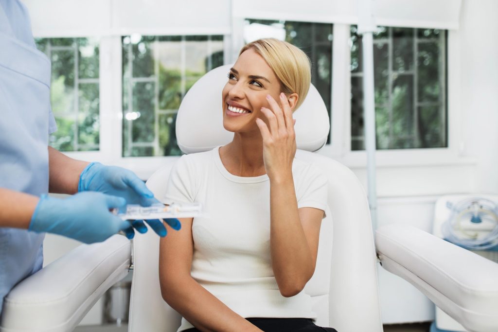 Smiling woman seated in an aesthetic treatment chair touching her cheek while a medical professional wearing blue gloves holds a syringe during a cosmetic consultation.
