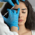 Medical professional wearing blue gloves administering a facial injection near a woman's forehead during a cosmetic injectable treatment.