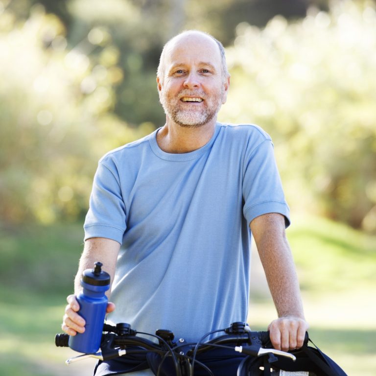 Smiling older man with short gray hair and a neatly trimmed beard wearing a light blue shirt outdoors, representing healthy aging and men's wellness.