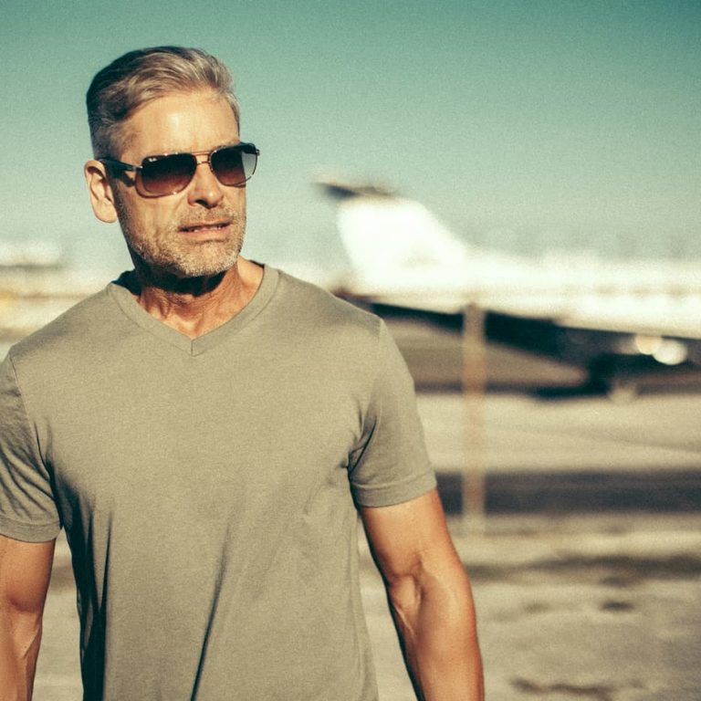 Confident middle-aged man with gray hair wearing sunglasses and a fitted gray T-shirt outdoors, representing men's health, vitality, and healthy aging.