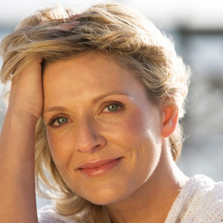 Close-up portrait of a smiling middle-aged blonde woman resting her hand against her forehead, representing women's hormonal wellness during perimenopause and menopause.