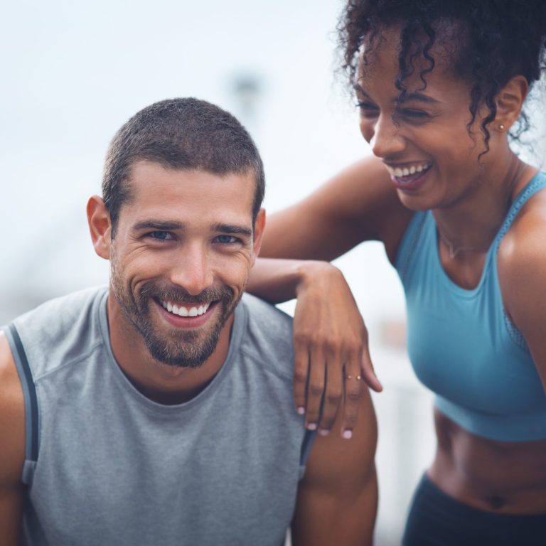 Smiling man in athletic clothing with a woman resting her arm on his shoulder, representing an active lifestyle, confidence, and support for men's health and wellness.